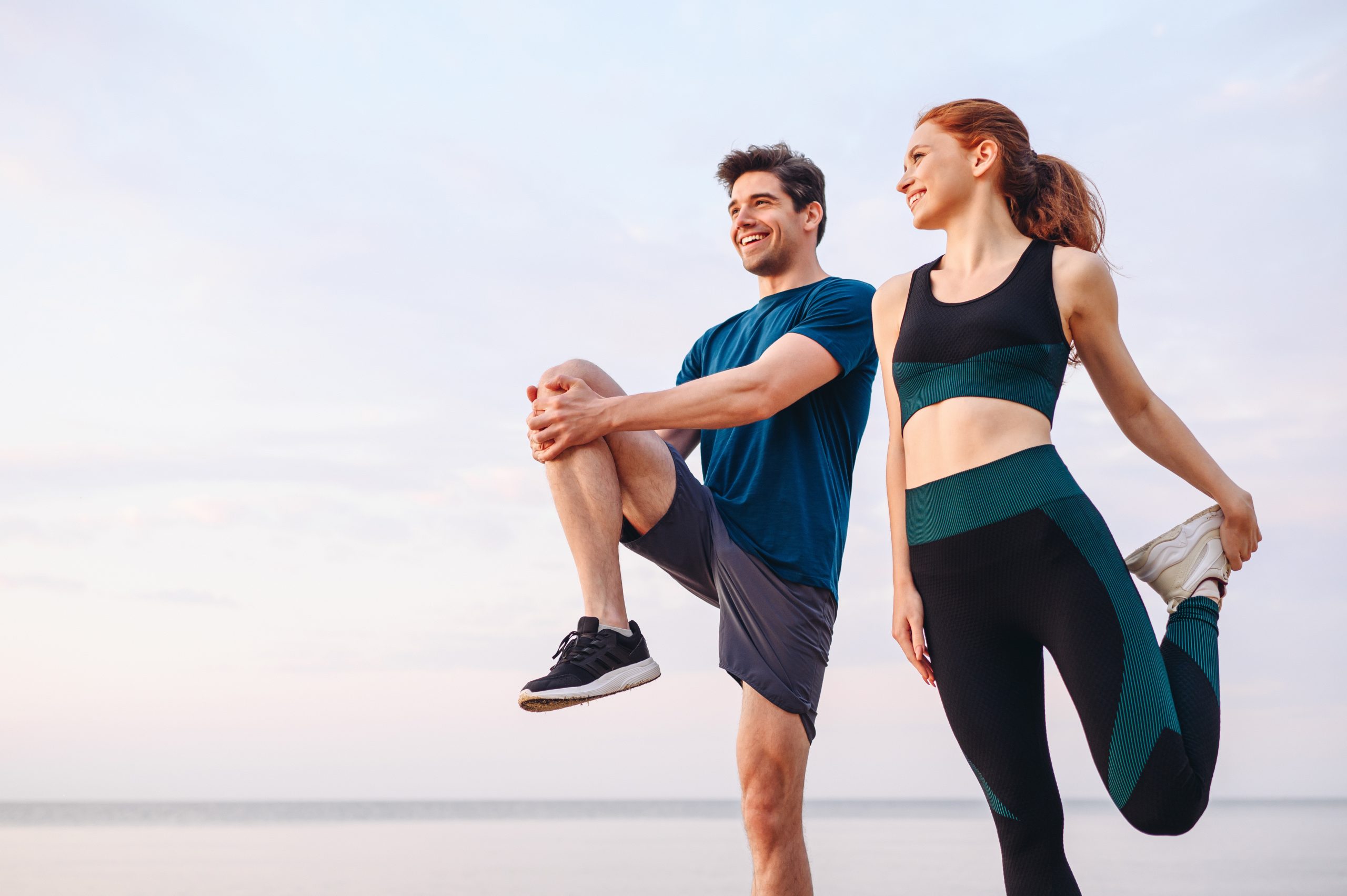 (alt text: homem e mulher, na praia, com roupa desportiva e a fazerem alongamentos para iniciarem exercício físico.)